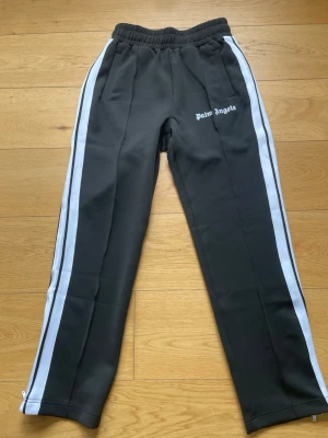 Palm Angels svarta mjukisbyxor - Svarta mjukisbyxor från Palm Angels med vita sidoränder och logga tryckt på vänster lår. Byxorna har resår i midjan, raka ben och dragkedjor vid bensluten. Tillverkade i ett glansigt, sportigt material som känns riktigt skönt och är helt nya 