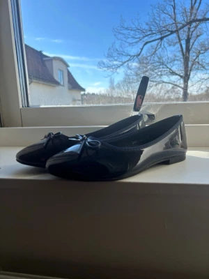 Ballerina skor  - Stilrena svarta ballerinaskor i glansigt lackmaterial med en söt rosett framtill. Skorna har rundad tå och låg sula, perfekta för en enkel men chic look. Passar till många olika outfits och ger en klassisk touch.