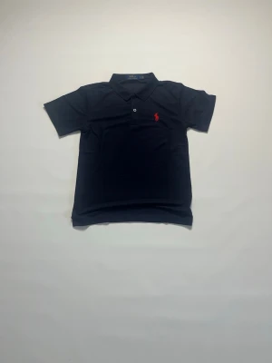 Marinblå pikétröja från Ralph Lauren storlek M - Klassisk Marinblå pikétröja från Ralph Lauren med korta ärmar och röd broderad logga på bröstet. Tröjan har krage och knappar framtill, perfekt för en clean och stilren look. Den är storlek M och är helt ny och aldrig buren