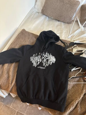 Antwan Hoodie - Svart hoodie med stor huva och coolt grafiskt tryck på bröstet i vitt och silver. Baksidan har ett stort gult tryck med texten 'Wanderland Antwan' och flera mindre detaljer. Klassisk känguruficka framtill och ribbade muddar. Perfekt för dig som gillar streetwear.