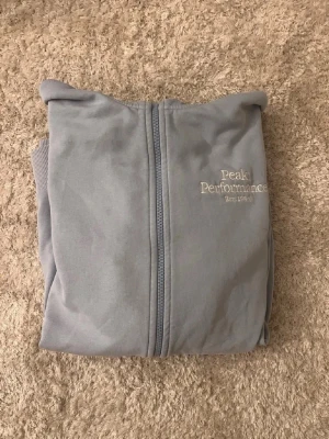 Ljusgrå hoodie från Peak Performance - Snygg ljusgrå hoodie från Peak Performance med dragkedja framtill och broderad logga på bröstet. Tillverkad i mjukt material som känns skönt mot huden. Perfekt för en avslappnad och sportig stil.