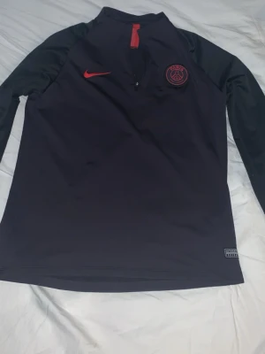 Svart PSG träningströja från Nike - Snygg svart långärmad träningströja från Nike med PSG-logga och röd Nike swoosh på bröstet. Tröjan har halv dragkedja framtill och är tillverkad i ett lätt, stretchigt material som passar perfekt för träning eller uppvärmning.