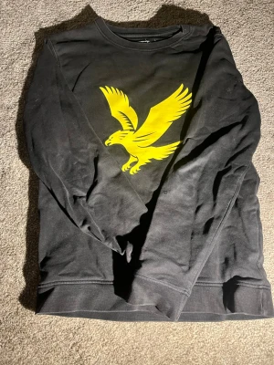 Svart sweatshirt med  Lyle and Scott logga på bröstet. Storlek junior large  - Cool svart sweatshirt med stor gul örn på bröstet. Tröjan har rund halsringning och långa ärmar. Perfekt för dig som gillar streetwear och vill sticka ut med en snygg grafisk detalj framtill. Storlek junior L
