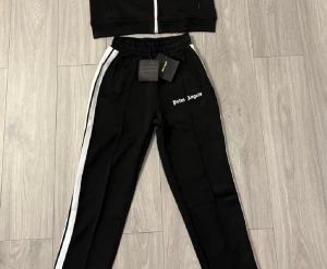 Palm Angels svart tracksuit set - Säljer ett svart tracksuit set från Palm Angels med vita ränder längs ärmar och ben. Setet består av en zip-jacka och byxor med resår i midjan. Båda delarna har Palm Angels-logga i vitt framtill. Sportig och trendig look. Hjälper en vän och sälja dom. 