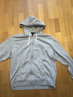 Grå zip-hoodie från H&M - Snygg grå hoodie från H&M i relaxed fit med dragkedja framtill, huva med vita snören och två fickor. Mjuk insida i bomullsblandning, perfekt för en chill och avslappnad stil.