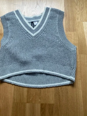 Grå stickad väst med v-ringning - Grå stickad väst med v-ringning och vita ränder vid hals och nederkant. Modellen är croppad och har en klassisk, ribbad kant. Perfekt att styla över en skjorta eller t-shirt för en trendig lager-på-lager-look.