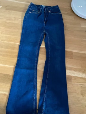 Blå utsvängda jeans med hög midja - Snygga blå jeans med utsvängda ben och hög midja. Klassisk femficksmodell med dragkedja och knapp framtill. Jeansen har en mörkblå tvätt och är tillverkade i denim. Perfekta för en trendig och avslappnad look. Den är klippt och sydd så skulle nog säga att den passar någon som är 155 mc. Den är liten i storlek så skulle passa någon som är XS
