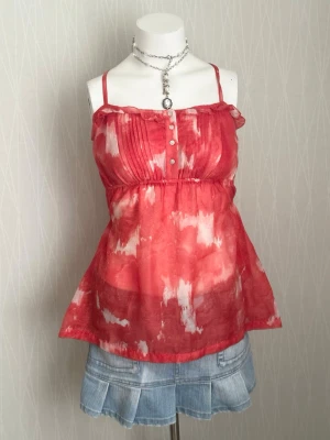 Röd och vit linnetopp med knappar - Snygg röd och vit linnetopp med tunna axelband och knappar framtill. Toppen har ett lätt utsvängt fall, rynk under bysten och ett abstrakt mönster. Perfekt till jeans eller kjol för en fräsch sommarlook.