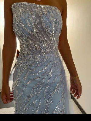 Ljusblå paljettklänning med slits - Säljer en elegant ljusblå långklänning med paljetter och transparenta detaljer. Klänningen är axelbandslös och har en snygg slits framtill. Perfekt för bal eller festliga tillfällen. Materialet känns lyxigt och skimrar vackert i ljuset. Passar 34/36