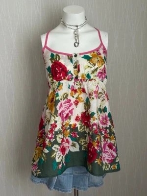 Blommig linnetopp med knappar - Söt blommig linnetopp med tunna axelband och knappar framtill. Toppen har rosa kantband och ett färgstarkt mönster i rött, grönt, gult och rosa på vit botten. Perfekt för varma dagar och ger en romantisk vibe.