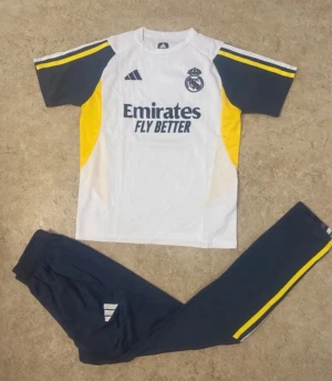 Real Madrid träningsset Adidas - Snyggt Real Madrid träningsset från Adidas med vit t-shirt och mörkblå byxor. Tröjan har gula och mörkblå detaljer, klubbmärke och Emirates Fly Better-tryck. Byxorna är mörkblå med gula ränder längs sidorna. Perfekt för fotbollsträning eller gymmet.