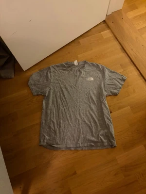 Grå T-shirt från The North Face - Säljer en grå T-shirt från The North Face med klassisk logga på bröstet och stor röd logga med texten 'Never Stop Exploring' på ryggen. T-shirten har rund hals och korta ärmar, perfekt för en avslappnad stil.