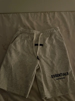 Grå Essentials Fear of God shorts - Snygga grå shorts från Essentials Fear of God med svart logga på vänster ben. De har elastisk midja med vit snörning och är tillverkade i mjukt bomullsmaterial. Perfekta för en avslappnad och trendig look.