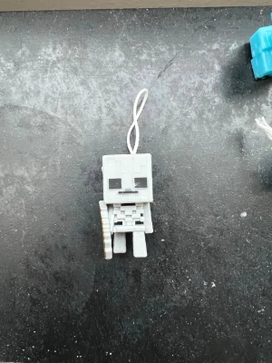 Kinder joy - Minecraft skelett nyckelring - Grå nyckelring i form av ett skelett från Minecraft. Tillverkad i plast med fyrkantig, pixlig design och ett vitt snöre för upphängning. Perfekt accessoar för att visa din gamingstil.