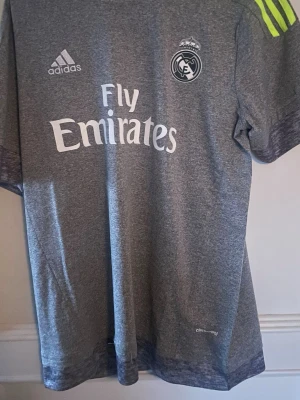 Real Madrid 15/16 Bale 11 - Snygg grå Real Madrid tröja från säsongen 15/16 med Real Madrid-logga och Fly Emirates-tryck på bröstet. Tröjan har korta ärmar och neongula ränder på axlarna. Tillverkad i lätt och ventilerande material med climacool-teknologi.