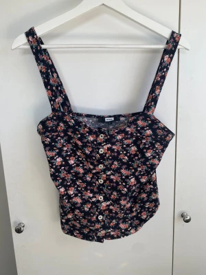 Blommig top  - Söt linnetopp från Bik Bok med blommigt mönster i rosa, grönt och vitt på mörkblå botten. Toppen har breda axelband och knappar framtill som detalj. Perfekt för varma dagar och enkel att styla med jeans eller kjol.