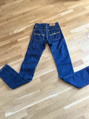  Nudie Jeans W25 L32 - Säljer ett par klassiska blå jeans från Nudie Jeans Co med raka ben och snygga kontrastsömmar. Jeansen har två bakfickor med Nudie Jeans karakteristiska broderi och är tillverkade i slitstark denim. Perfekta för en avslappnad och trendig look.
