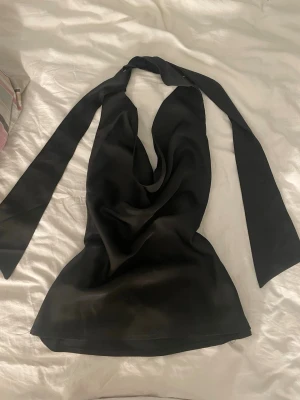 Svart omlottopp i satin - Snygg svart omlottopp i satin med halterneck och långa knytband.storlek L men passar mig som har storlek S. Toppen har en draperad framsida som ger en cool look och passar perfekt till jeans eller kjol. Materialet är glansigt och mjukt vilket gör den extra skön att bära.