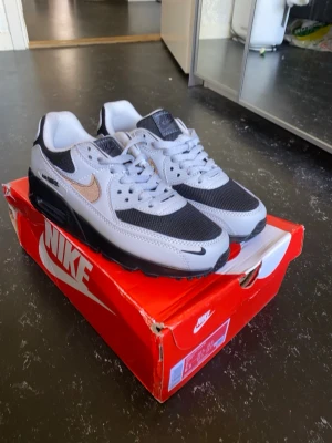 Nike Air Max 90 grå och svarta  - Nike Air Max 90 sneakers i grått och svart med silvrig swoosh. Skorna har klassisk chunky sula, mesh och läderdetaljer samt synlig Air Max-dämpning. Perfekt för dig som gillar streetwear och sportig stil.