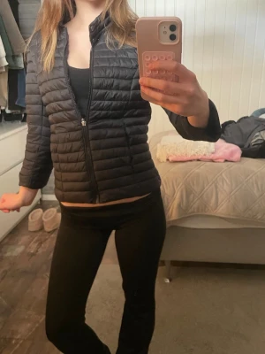 Herno liknande jacka - Säljer en svart pufferjacka med quiltad design och hög krage. Jackan har dragkedja framtill och tryckknappar vid ärmsluten. Perfekt för kalla dagar och enkel att matcha med olika outfits.