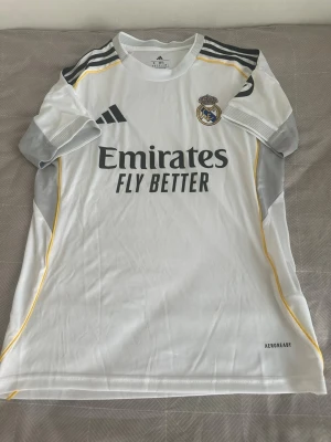 Real Madrid matchtröja Adidas vit - Snygg vit Real Madrid matchtröja från Adidas med svarta och gula detaljer. Tröjan har klubbmärke, sponsortryck och AEROREADY-material som andas. Grå partier på ärmar och sidor samt HP-logga på ena ärmen. Perfekt för fotboll eller träning. Använd ett par få gånger 😁