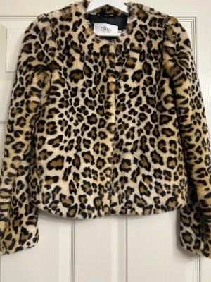 Leopardmönstrad fuskpälsjacka Stand - Snygg leopardmönstrad fuskpälsjacka från Stand. Jackan har rund halsringning, raka ärmar och dold knäppning framtill. Mjuk och fluffig känsla, perfekt för att sticka ut i vinter. Djurmönstret går i beige, brunt och svart.