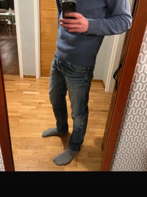 Nudie Jeans - Säljer ett par klassiska blå jeans från Nudie Jeans i modellen straight fit. Jeansen har snygga slitningar och kontrastsömmar, samt Nudie-loggan broderad på bakfickorna. Tillverkade i slitstark bomull med femficksdesign och normal passform. Sitter som 30/32