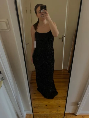 Balklänning - Snygg svart långklänning med glitter och smala axelband. Klänningen har öppen rygg med knytning. Endast testad och har alla lappar kvar, säljer då den va för stor🥰