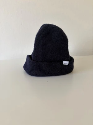 Mörkblå merinoull mössa Les Deux - Snygg mörkblå ribbstickad mössa från Les Deux med uppvikt kant och liten vit logolapp på sidan. Klassisk modell som sitter skönt på huvudet och passar perfekt till kyliga dagar. Tillverkad i merinoull