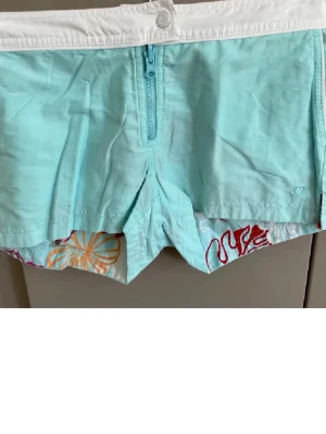 Turkosa badshorts med vit linning - Säljer ett par turkosa badshorts med vit linning och dragkedja framtill. Insidan har färgglatt blommigt mönster i orange och rött. Shortsen har kort modell och är tillverkade i lätt, snabbtorkande material. Perfekta för stranden eller poolen! Tvåsidig. 