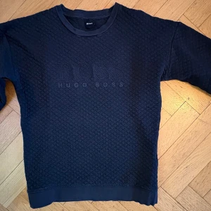 Svart quiltad tröja från Hugo Boss - Säljer en svart tröja från Hugo Boss med quiltad struktur och diskret logga broderad på bröstet. Tröjan har rund halsringning, ribbade muddar och en clean look som passar perfekt till streetwear-stilen.