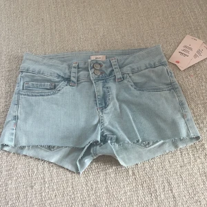 Ljusblå jeansshorts från Nelly - Helt nya snygga ljusblå jeansshorts från Nelly med råa kanter och dubbla knappar framtill. Shortsen har klassiska fickor och coola bakfickor med rosa broderad detalj. Perfekta för varma dagar och enkel att matcha med allt.