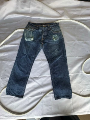 Nudie jeans med slitningar och feta fades - Snygga blå jeans med raka ben och coola slitningar framtill vid fickorna. Klassisk femficksmodell med orangea sömmar och knappgylf. Perfekta för en avslappnad streetstil. Materialet är kraftig denim i bomull. Mobil och AirPods fades. W34 L30