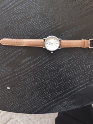 Klocka från Karl-Axel stockholm med läderband - Stilren klocka från Karl-Leimon med rund silverfärgad boett och ljus urtavla. Klockan har ett klassiskt brunt läderarmband och minimalistisk design med tydliga indexmarkeringar. Perfekt accessoar för en clean look.