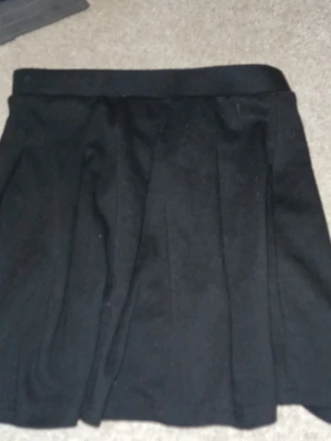 Svart kortkjol med veck - En svart kortkjol med klassiska veck och bred linning. Kjolen har en enkel och stilren design som passar till många olika outfits. Perfekt för dig som gillar en tidlös look.