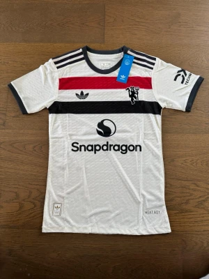Manchester United bortatröja Adidas - Snygg Manchester United bortatröja från Adidas med svart och röd rand över bröstet, svart logga och sponsortryck. Nummer 8 och B. Fernandes på ryggen. Tillverkad i lätt funktionsmaterial med HEAT.RDY-teknologi. Perfekt för fotbollsträning eller match.