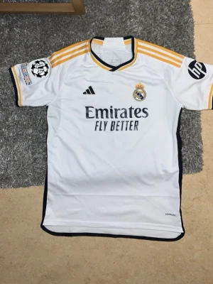 L. Äkta Real Madrid tröja 2024 - Hellt ny den har vart hemma i 2 år uttan använda köpte den till en vän som present gav den till honom han hade XL visste inte trodde han hade L såklart tog av prislappen för det är en present därför kan inte returnera tillbaka och valde sälja den här en vit Real Madrid matchtröja från Adidas med Vini Jr och nummer 7 på ryggen. Tröjan har gula och blå detaljer , klubbmärke och sponsortryck framtill samt Champions League-märke på ärmen. Perfekt för fotbollsfans!