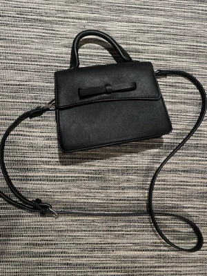 Svart handväska med rosett - Stilren svart handväska i slät konstläder med dekorativ rosett framtill. Väskan har både handtag och en lång justerbar axelrem, perfekt för att bära på olika sätt. Enkel och elegant design som passar till många outfits. Har nästan aldrig används.