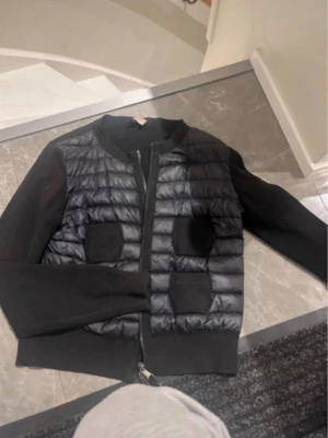 Svart cardiganjacka från Moncler - Snygg svart cardiganjacka från Moncler med quiltad framsida och två fickor. Jackan har stickade ärmar och rygg, dragkedja framtill och ribbade muddar. Perfekt för lager på lager och ger en sportig men ändå stilren look. Den är i storlek M men den har krympt i tvätten så passar S också