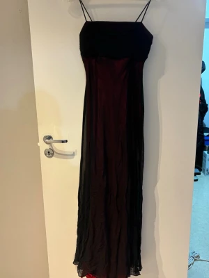 Svart och röd långklänning med tunna band - Elegant långklänning med tunna axelband i svart och rött. Klänningen har en figurnära överdel och ett långt, flytande kjolparti med svart transparent tyg över röd bas. Perfekt för en snygg och stilren look.