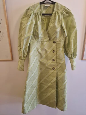 Ljusgrön omlottklänning med puffärm - Snygg ljusgrön långklänning från Ganni med omlott-design och diagonala ränder. Klänningen har puffiga långärmar, djup v-ringning och dekorativa knappar framtill. Tillverkad i ett glansigt material som ger en lyxig känsla.