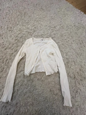 Vit ribbad kofta från Kappahl - Säljer en vit ribbad kofta från Kappahl med långa ärmar och rund halsringning. Koftan har knäppning framtill med vita knappar och är i en croppad modell. Perfekt att slänga över en topp för en chill look.