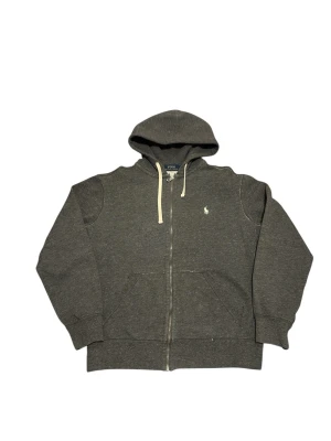 Mörkgrå hoodie från Polo Ralph Lauren - Mörkgrå hoodie med dragkedja från Polo Ralph Lauren. Klassisk design med vit broderad logga på bröstet, huva med vita snören och stora fickor framtill. Tillverkad i mjukt bomullsmaterial som är perfekt för en avslappnad stil.                                              📍Storlek:M  💪🏽Skick:Ny utan prislapp   📐Mått:Längd:58:cm:Bred:50:cm:  📎Material:86%Bomull:14%polyester  📦Skickar alltid inom 48h  ✅Äkthetsgaranti   