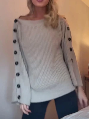 Beige stickad tröja med knappar - Trendig beige stickad tröja med bred ribbstickning och svarta dekorativa knappar längs ärmarna. Tröjan har en avslappnad passform, rund halsringning och vida ärmar. Perfekt för lager-på-lager och en cool vardagslook.