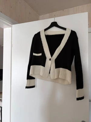 Svartvit ribbstickad kofta - Trendig svart och vit ribbstickad kofta med djup V-ringning och en knapp framtill. Koftan har kontrastfärgade muddar, en liten ficka på bröstet och är något croppad i modellen. Perfekt för lager-på-lager och snygg till både jeans och kjol.