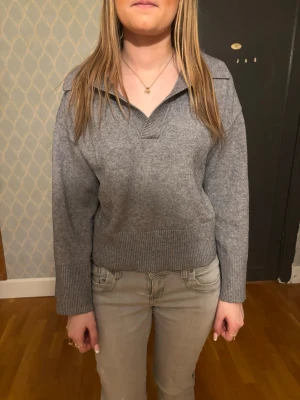 Grå stickad tröja med krage - Mysig grå stickad tröja med bred ribbad mudd och krage. Tröjan har en avslappnad passform, långa ärmar och en djupare v-ringning utan knappar. Perfekt till jeans för en chill look.