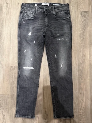 Replay Anbass Mörkgrå Jeans W32/L30 Aged 10 Years - Replay Anbass jeans i mörkgrå tvätt med snygga slitningar och “Aged 10 Years”-look, en clean och tidlös modell som funkar till det mesta, storlek W32/L30, passform slim/regular beroende på kroppstyp, midja 42,5 cm, ytterbenslängd 99,5 cm, benöppning 17 cm, modellen är 185 cm och väger 65 kg, i fint skick med naturligt slitage som ger jeansen sin stil.