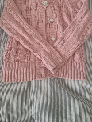 Rosa kabelstickad kofta - Mysig rosa kabelstickad kofta med stora vita knappar framtill. Koftan har ribbade muddar vid ärmslut och nederkant samt ett klassiskt kabelmönster. Superfin rosa färg, perfekt nu i vår och sommar!🌸 Storlek m, men skulle snarare säga att den sitter som s.