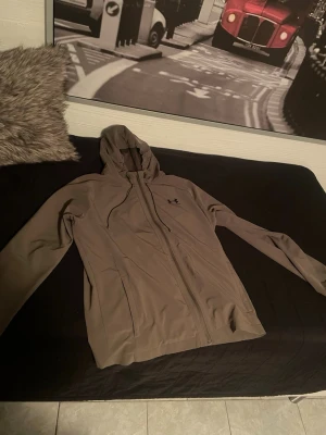 Beige vindjacka från Under Armour - Snygg beige vindjacka från Under Armour med huva och dragkedja framtill. Jackan har två sidofickor och en minimalistisk design med diskret logga på bröstet. Perfekt för blåsiga dagar och enkel att matcha med olika outfits, byxor finns också men hela priset är då 1000, jackan är köpt för 1000 o byxorna 600