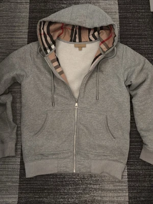 Grå zip hoodie från Burberry - Snygg grå hoodie från Burberry med dragkedja, klassiskt rutigt foder i huvan och broderad logga på bröstet. Mjuk bomullsblandning, två fickor fram och ribbade muddar. Perfekt för en chill och stilren look.